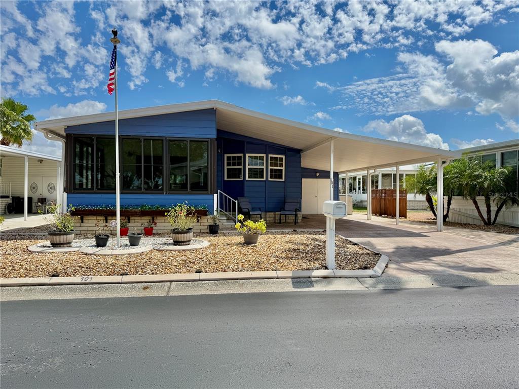 A blue single-story manufactured home features a large carport, a flag pole, and gravel landscaping — 101 SAINT GEORGE COURT, APOLLO BEACH, FL