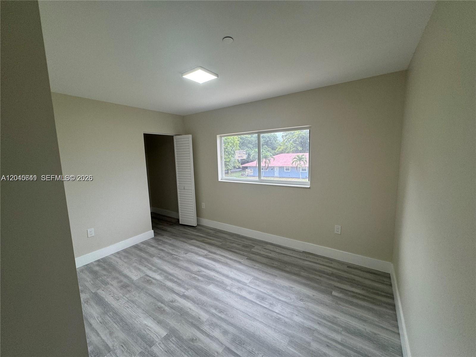 An empty bedroom features light-colored walls, gray wood-look flooring, and a window overlooking a house — 205 NW 159th St, Miami FL 33169, MIAMI, FL