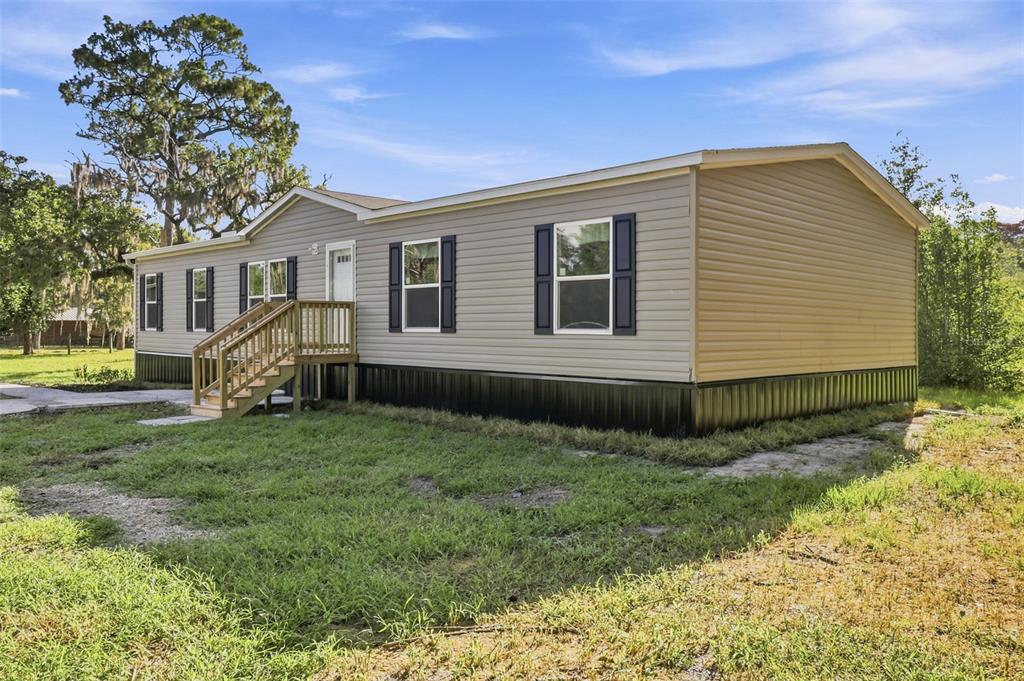 A tan manufactured home with black skirting and shutters sits on a grassy lot — 18422 MOORHAVEN DRIVE, SPRING HILL, FL