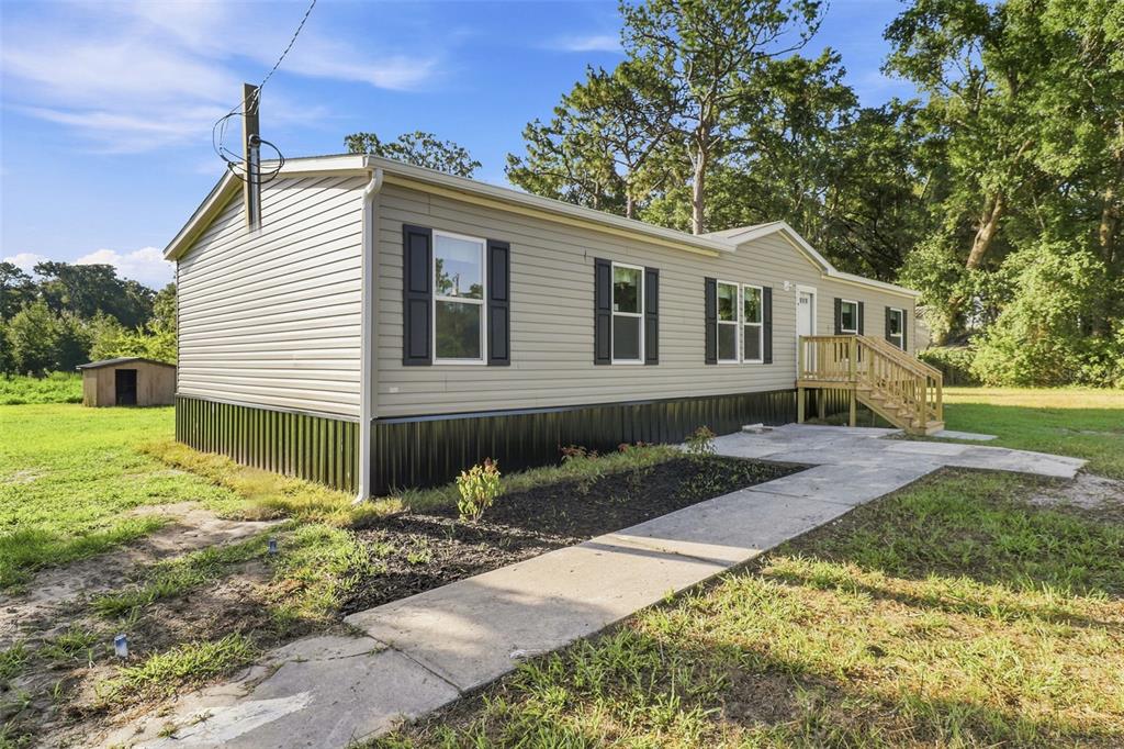 A light-colored manufactured home with black skirting and a concrete walkway sits on a lawn — 18422 MOORHAVEN DRIVE, SPRING HILL, FL