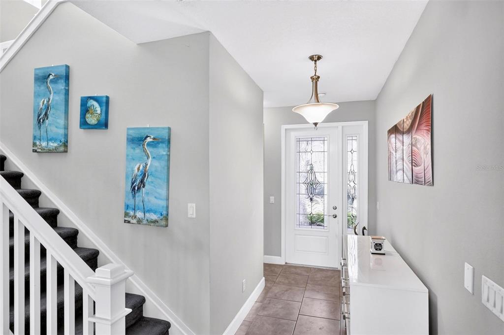 A bright entryway features a staircase with dark carpet, tiled floors, and coastal wall art — 3073 MOUNTAIN SPRUCE TERRACE, WESLEY CHAPEL, FL
