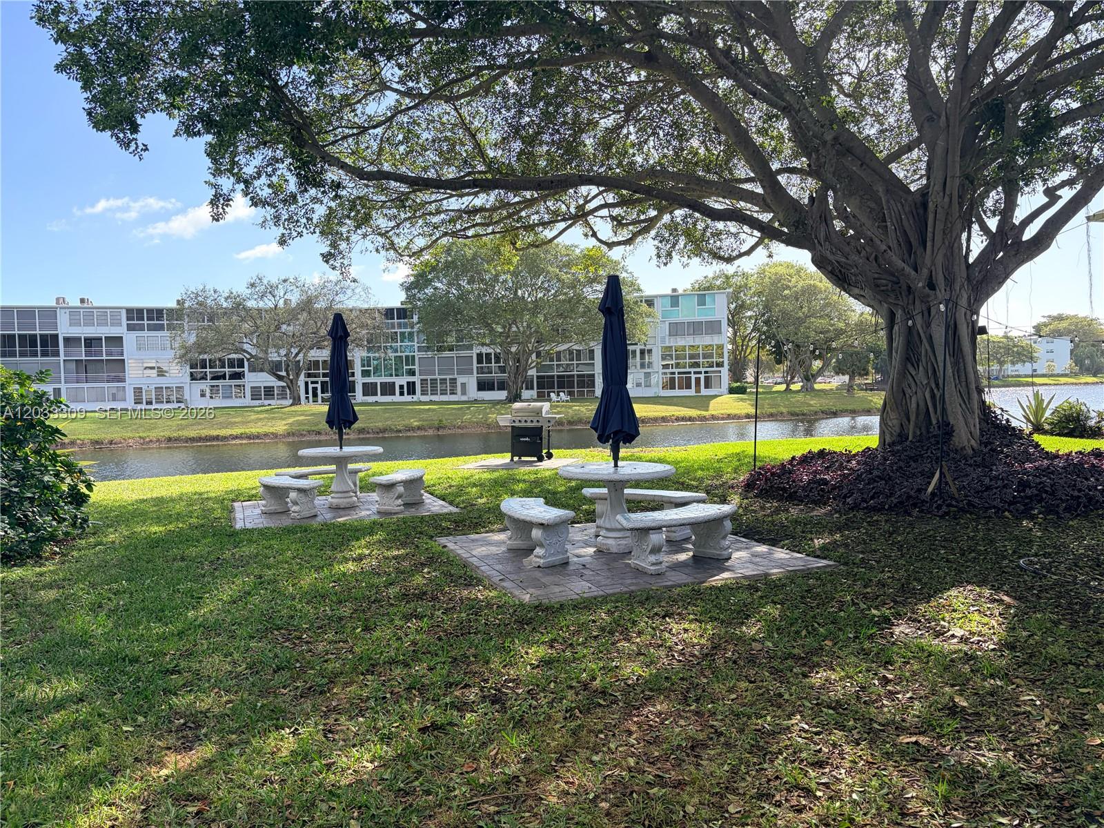 Two stone picnic tables with umbrellas sit on a lawn near a canal and tree — 178 Durham C # 178, Deerfield Beach FL 33442, DEERFIELD BEACH, FL