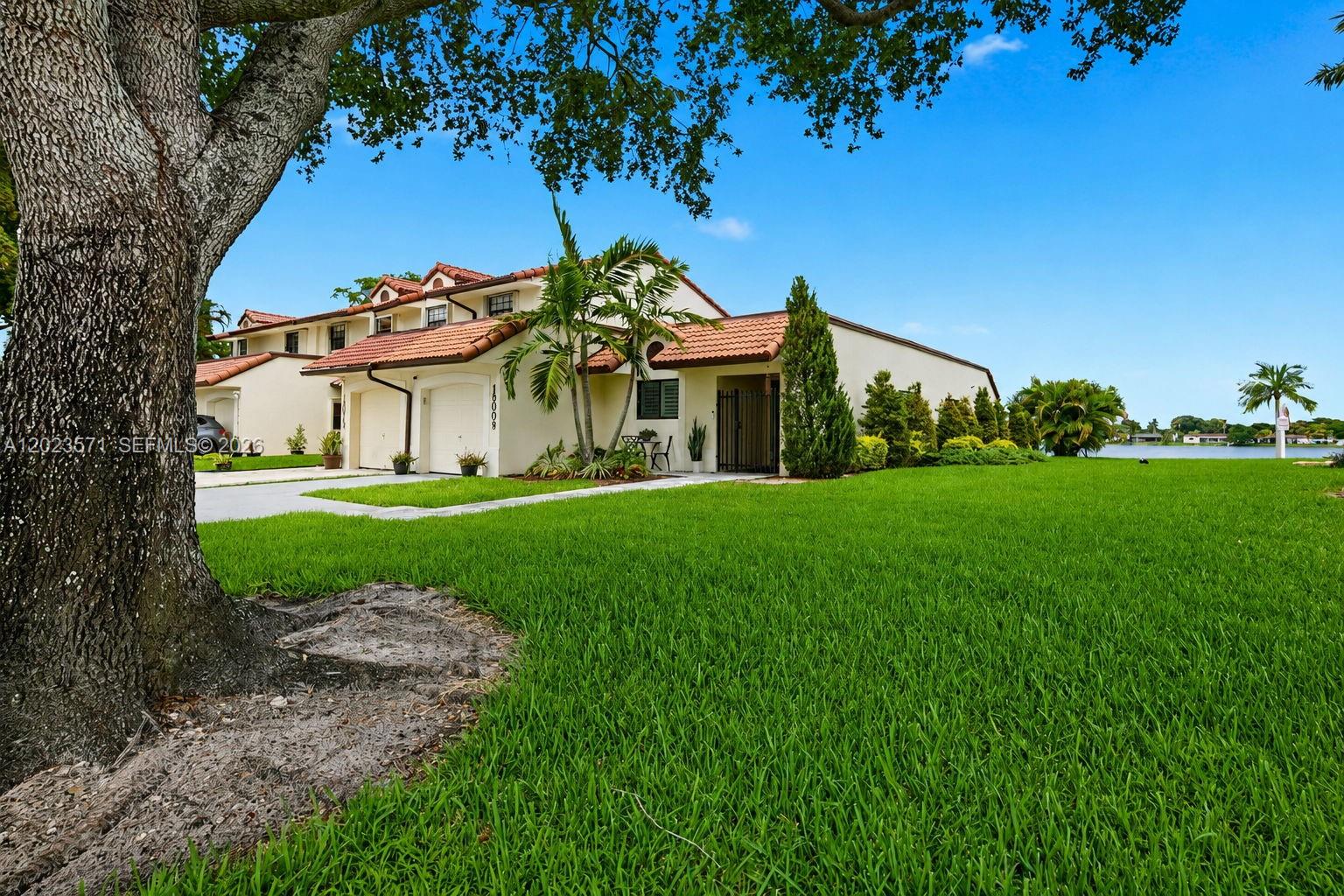 A single-story townhouse with a tile roof, garage, and palm trees sits near a lake — 19009 NW 64th Ct, Hialeah FL 33015, HIALEAH, FL