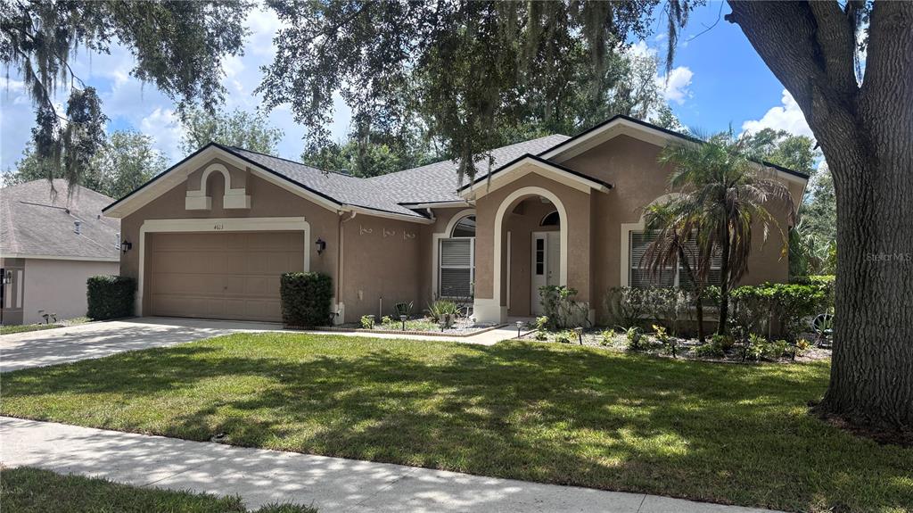 A single-story tan house with a two-car garage and a covered entryway under large trees — 4613 RIVER OVERLOOK DRIVE, VALRICO, FL