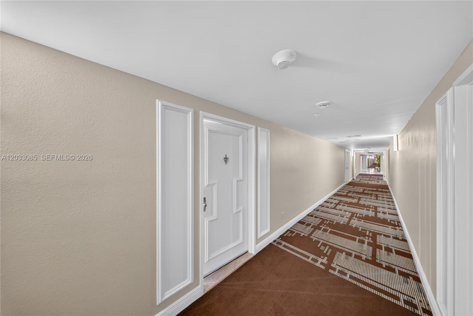A long hotel-style hallway features beige walls, white trim, and a patterned brown carpet — 5600 Collins Ave # 7W, Miami Beach FL 33140, MIAMI BEACH, FL