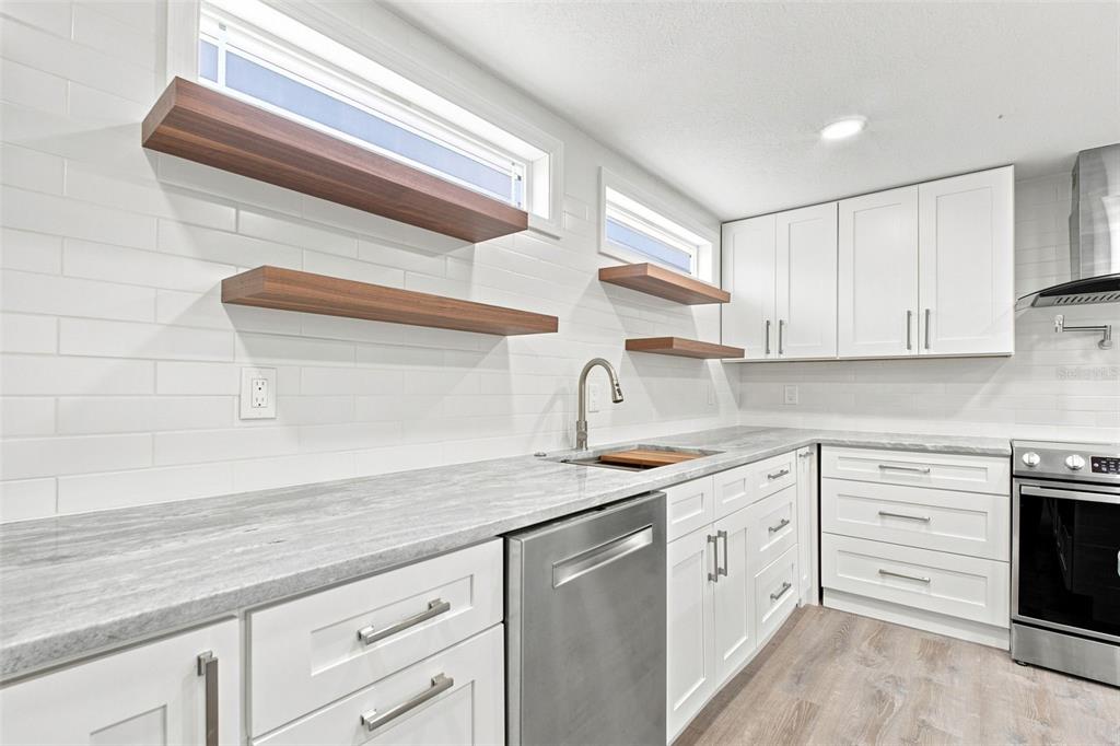 A modern kitchen features white cabinets, gray stone countertops, wood floating shelves, and stainless appliances — 163 DOLPHIN DRIVE E, OLDSMAR, FL