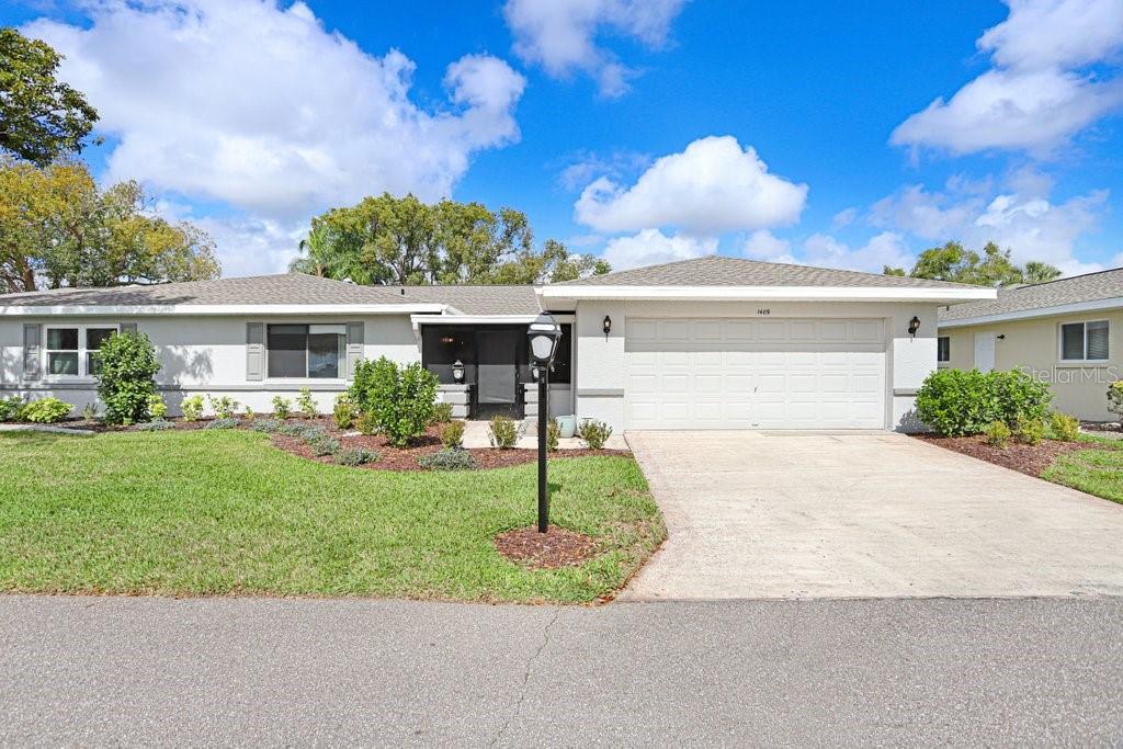 A single-story house with a light-colored exterior, a two-car garage, and a landscaped front yard — 1409 INGRAM DRIVE UNIT 75, SUN CITY CENTER, FL