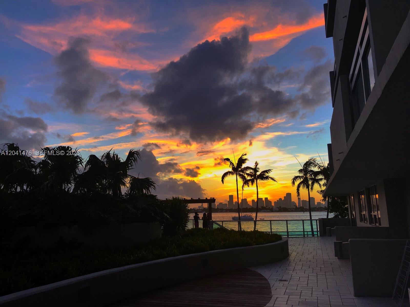A sunset view over a bay with palm trees, a skyline, and a paved patio — MIAMI BEACH, FL