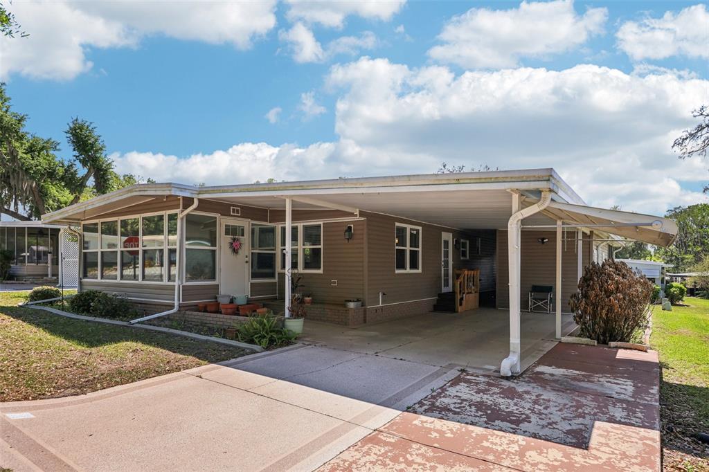 A single-story manufactured home features tan siding, a large carport, and a concrete driveway — 105 ORCHID WAY, LEESBURG, FL