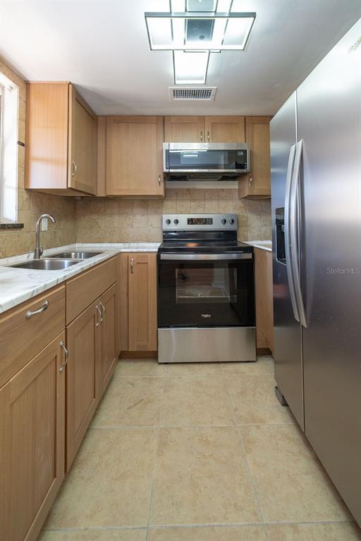 A galley kitchen features light wood cabinets, stainless steel appliances, and beige tile flooring — 4807 MARINE PARKWAY UNIT 103, NEW PORT RICHEY, FL