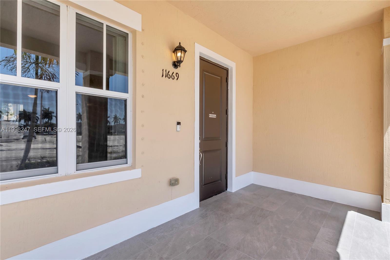 A covered entryway features a brown door, a wall-mounted light, and tiled flooring — 11669 SW 247th St, Homestead FL 33032, HOMESTEAD, FL