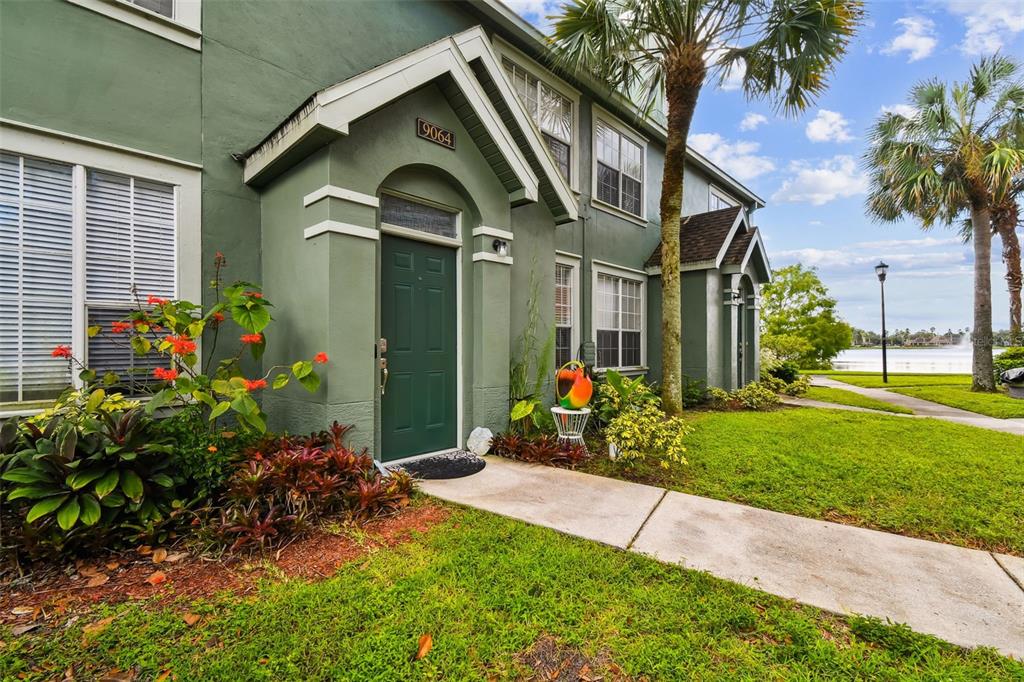 A green two-story townhouse exterior features a dark door, white trim, and a nearby lake — 9064 LAKE CHASE ISLAND WAY, TAMPA, FL