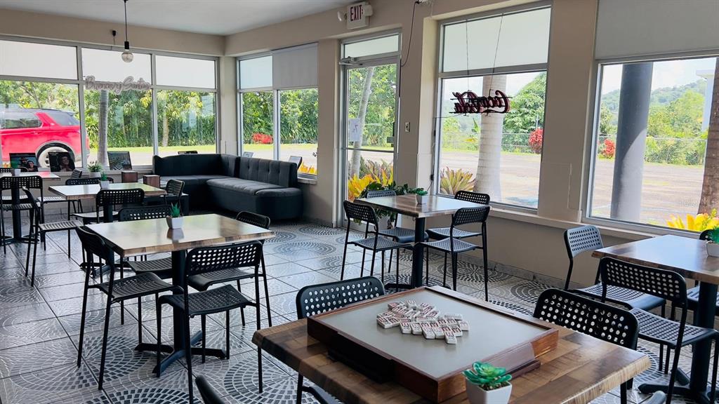 A bright restaurant dining area features multiple tables, chairs, a corner sofa, and large windows — Km 2.4 CARR 402, ANASCO, PR