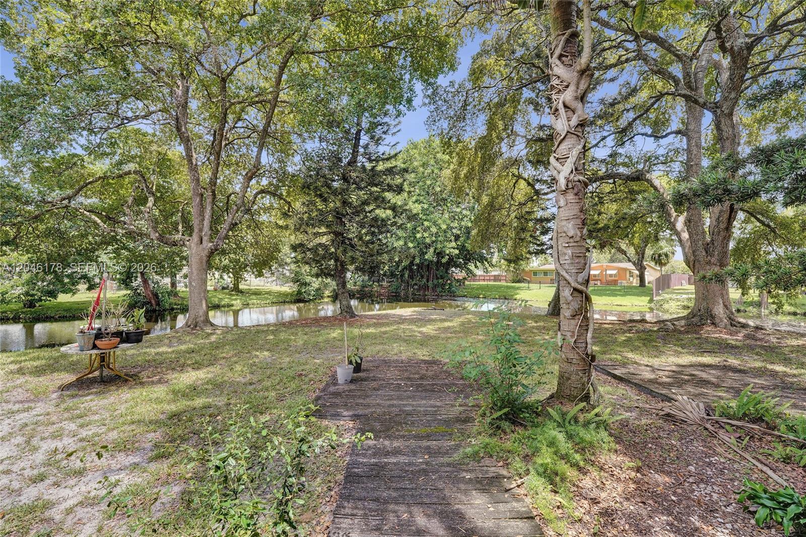 A grassy backyard features a wooden walkway leading toward a small pond and mature trees — 9120 SW 49th St, Cooper City FL 33328, COOPER CITY, FL