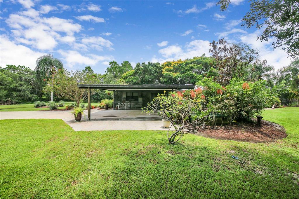 A covered concrete patio sits in a grassy backyard surrounded by trees and landscaping — 6168 ARBOREA DRIVE, INDIAN LAKE ESTATES, FL