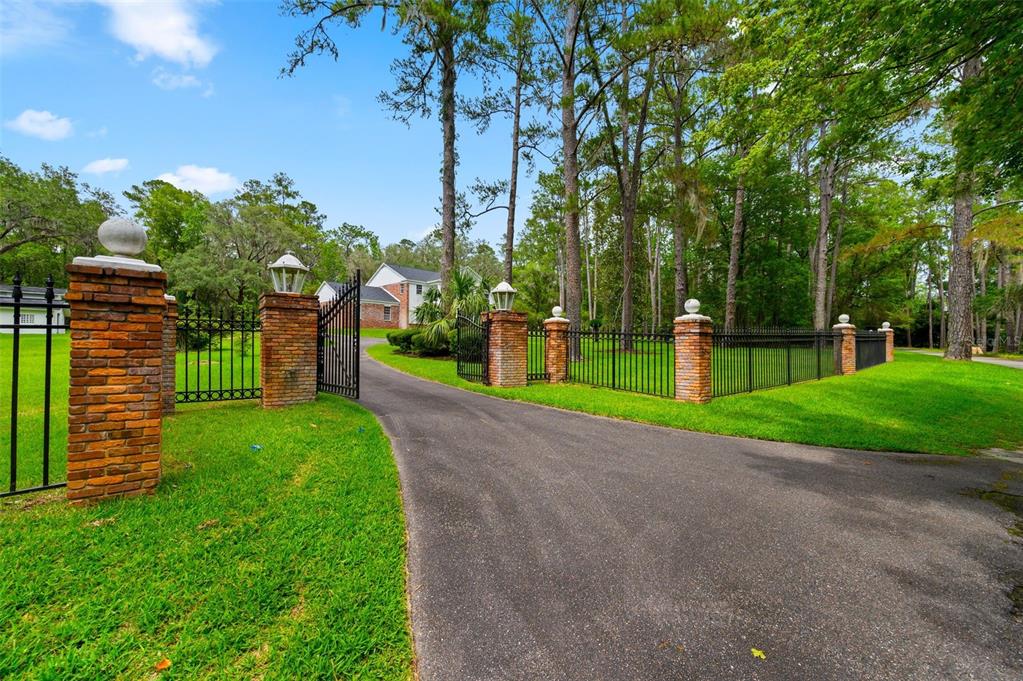 A paved driveway leads through an open wrought iron gate flanked by brick pillars — 7216 NW 20TH PLACE, GAINESVILLE, FL