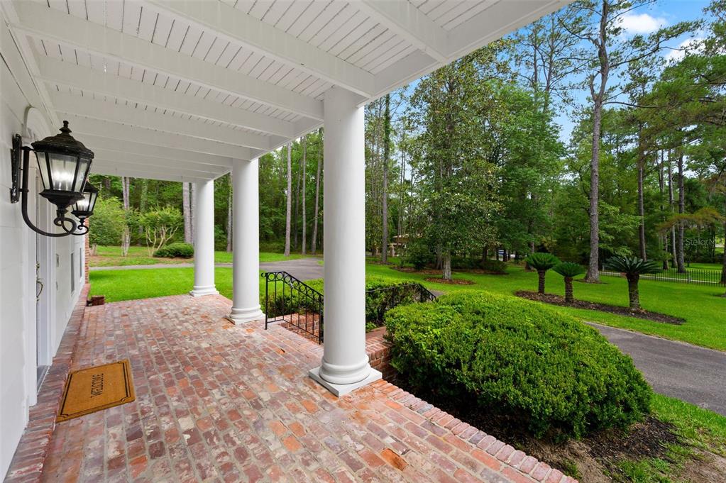 A covered brick porch with white columns overlooks a grassy yard and wooded area — 7216 NW 20TH PLACE, GAINESVILLE, FL