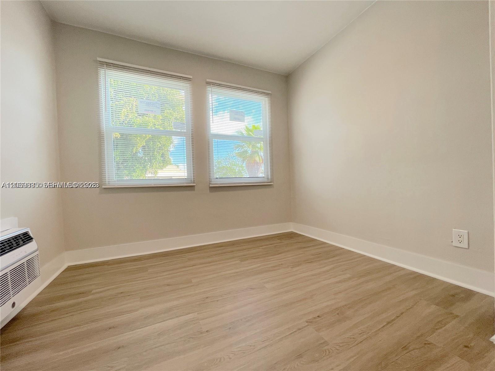 An empty room features light-colored walls, wood-look flooring, two windows with blinds, and an air conditioner — 1836 Dewey St # 2, Hollywood FL 33020, HOLLYWOOD, FL