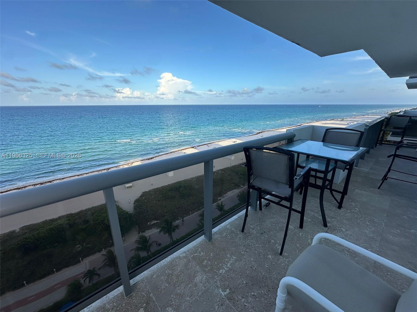 A balcony with a glass table and chairs overlooks the ocean and a sandy beach — 6039 Collins Ave # 1517, Miami Beach FL 33140, MIAMI BEACH, FL