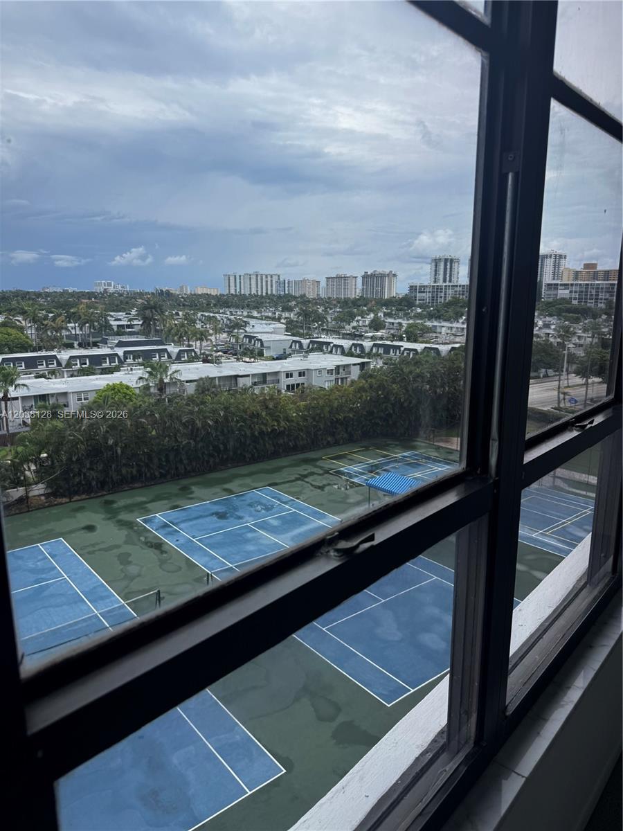 A high-angle view through a window shows tennis courts below and a distant cityscape — HALLANDALE BEACH, FL