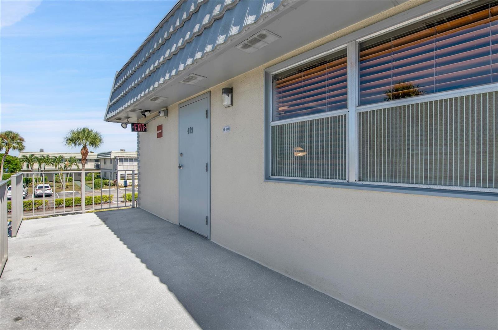 An exterior view of a building walkway featuring a gray door, window, and metal railing — 409 Brittany I # 409, Delray Beach FL 33446, DELRAY BEACH, FL