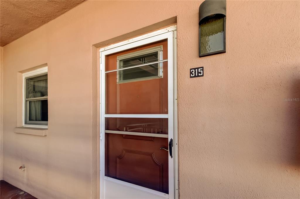 A peach-colored exterior wall features a front door with a screen, a window, and house number 315 — 5521 80TH STREET N UNIT 315, ST PETERSBURG, FL