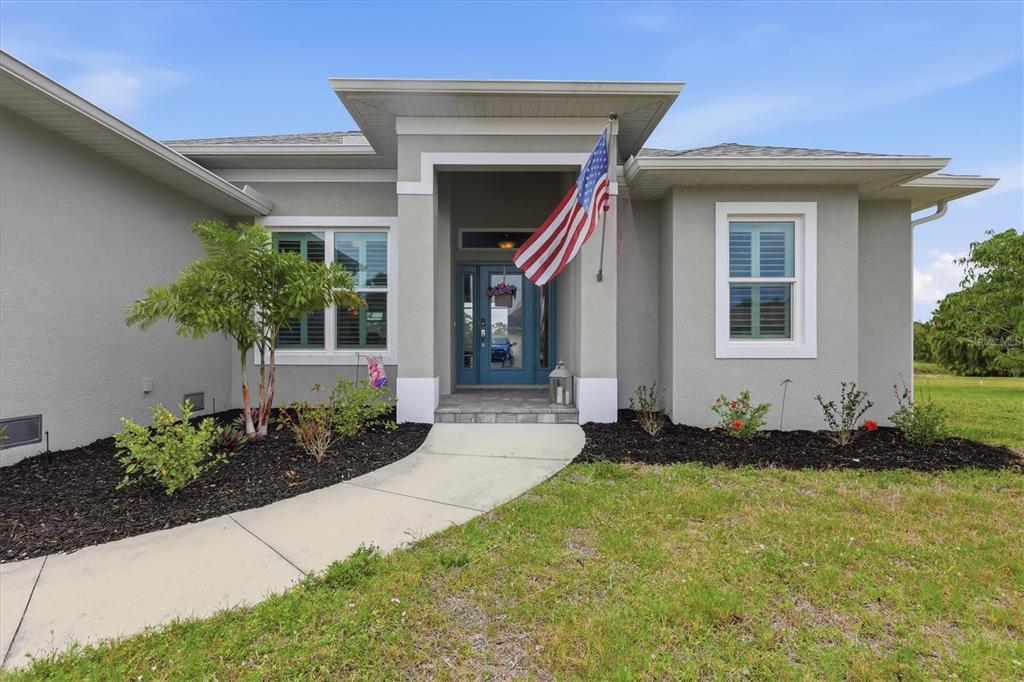 A single-story house exterior features a gray stucco facade, a blue front door, and an American flag — 48 LONG MEADOW LANE, ROTONDA WEST, FL