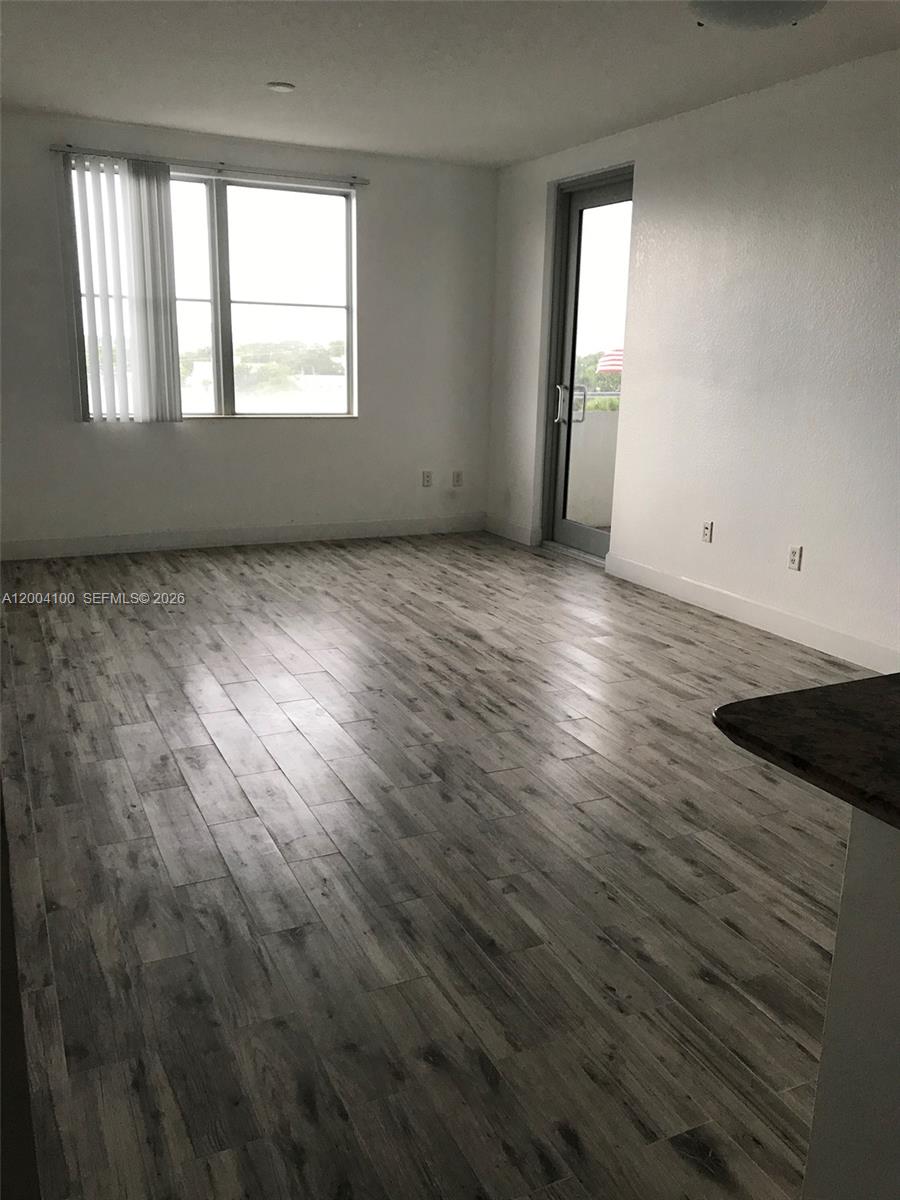 An empty room features wood-look tile flooring, white walls, a large window, and a balcony door — 1830 Radius Dr # 407, Hollywood FL 33020, HOLLYWOOD, FL