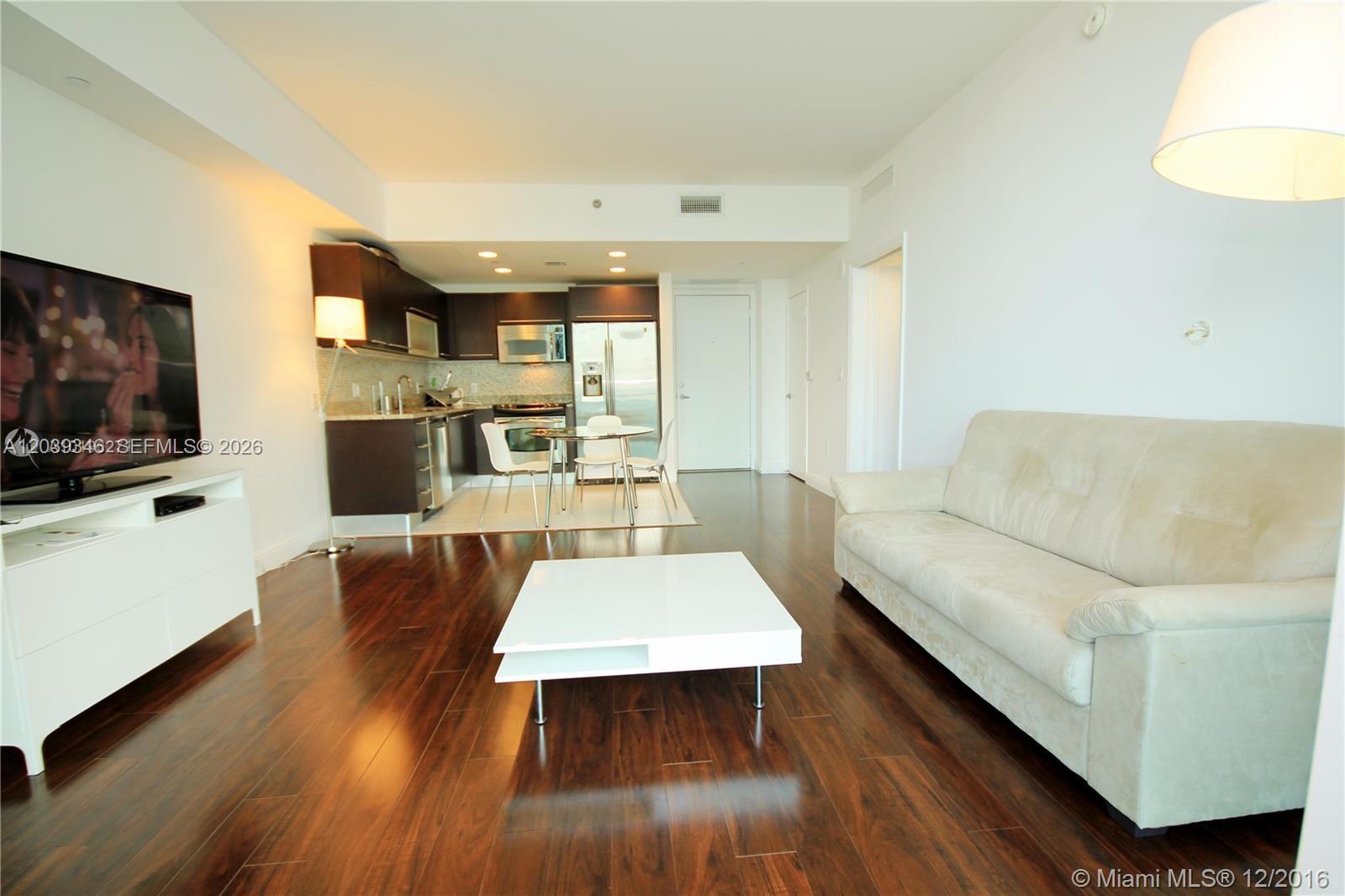 A living room with dark wood floors features a white sofa, coffee table, and kitchen — MIAMI, FL