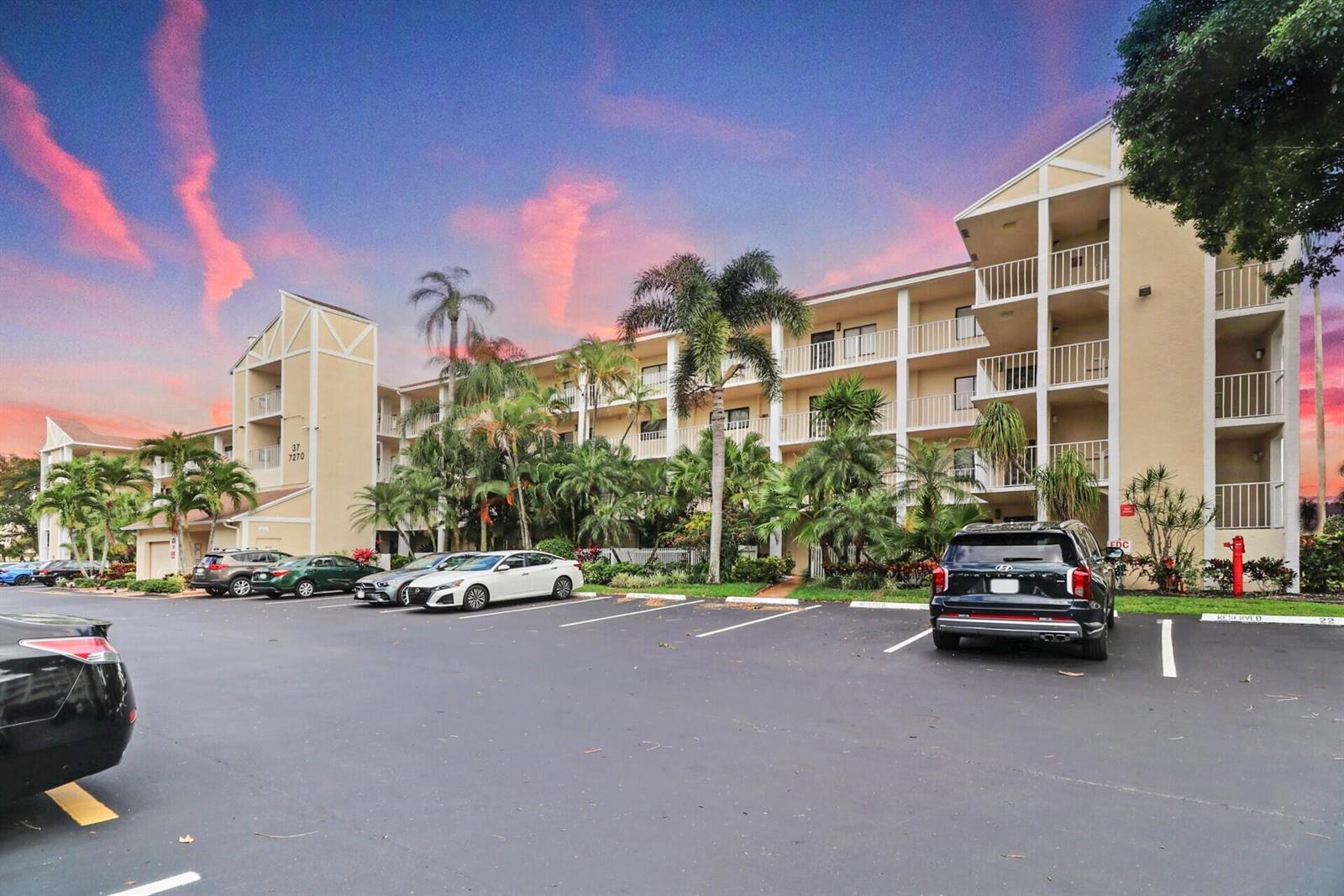 A multi-story apartment building with balconies and palm trees sits behind a paved parking lot — 7270 Ashford Place # 106, Delray Beach FL 33446, DELRAY BEACH, FL