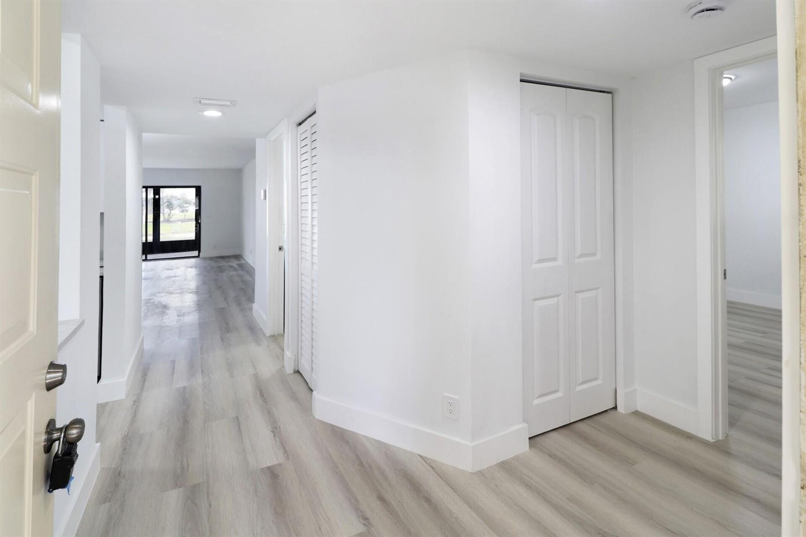 A hallway with light wood-look flooring leads to a bright living area with a glass door — 7270 Ashford Place # 106, Delray Beach FL 33446, DELRAY BEACH, FL