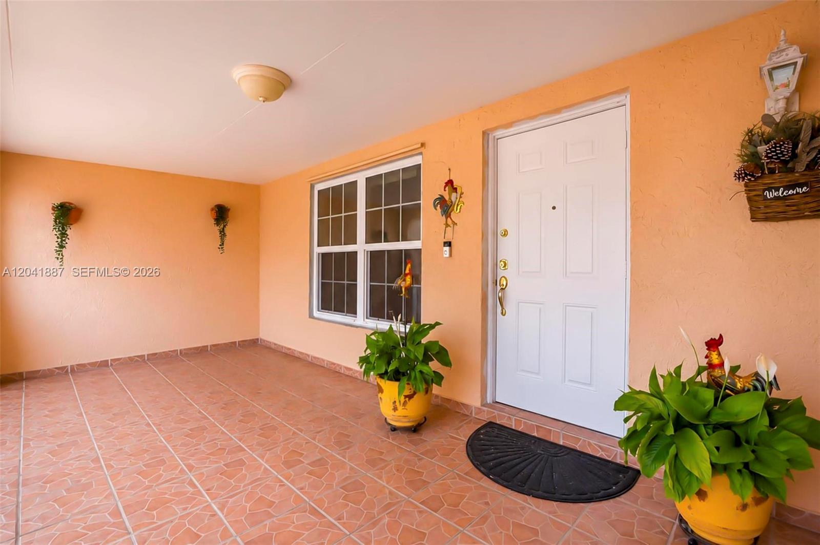 A covered porch features peach-colored walls, tiled flooring, a white front door, and potted plants — 19240 NW 60th Ct, Hialeah FL 33015, HIALEAH, FL