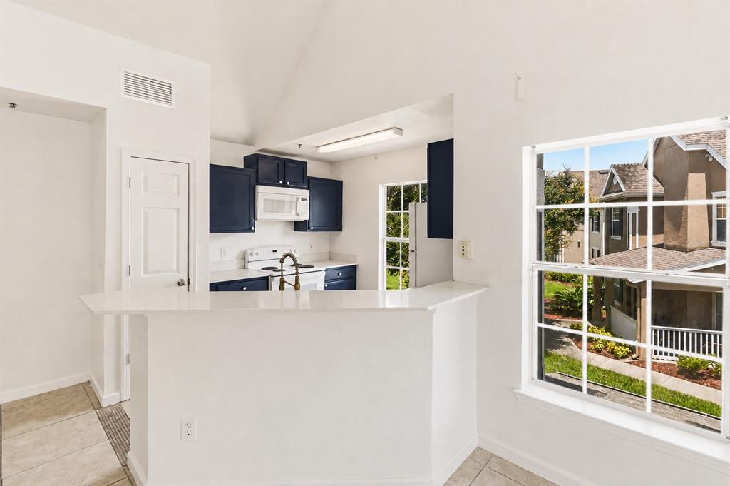 A kitchen with dark blue cabinets and white countertops overlooks a dining area with windows — 9157 LEE VISTA BOULEVARD UNIT 205, ORLANDO, FL