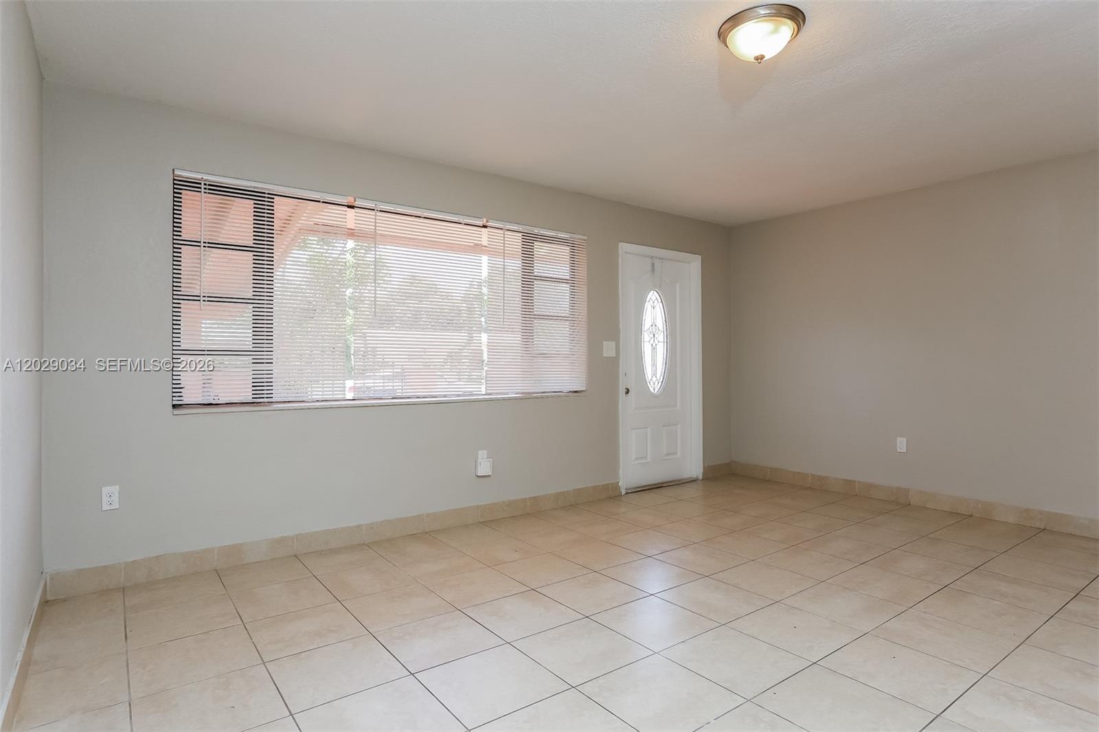 An empty room features beige tile flooring, light gray walls, a large window, and a door — 731 NW 175th Dr, Miami Gardens FL 33169, MIAMI GARDENS, FL