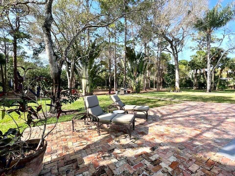 Two lounge chairs sit on a brick patio overlooking a grassy yard with tall trees — 14256 Greentree Trail, Wellington FL 33414, WELLINGTON, FL