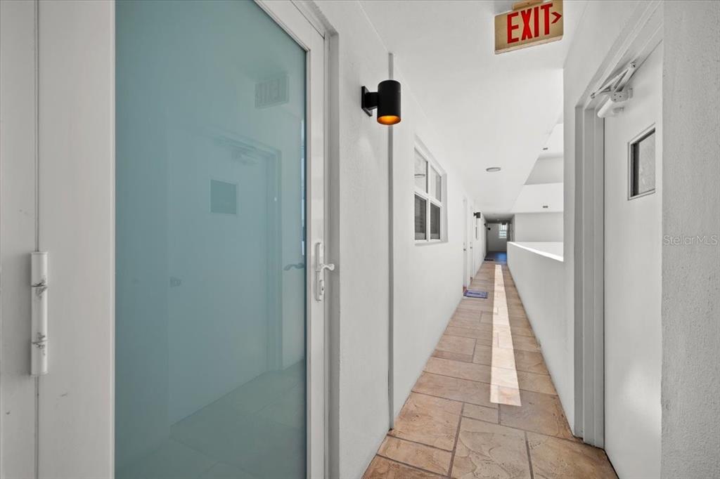 A tiled outdoor hallway features white walls, a frosted glass door, and an exit sign — 1630 N OCEAN BLVD UNIT 1011, POMPANO BEACH, FL