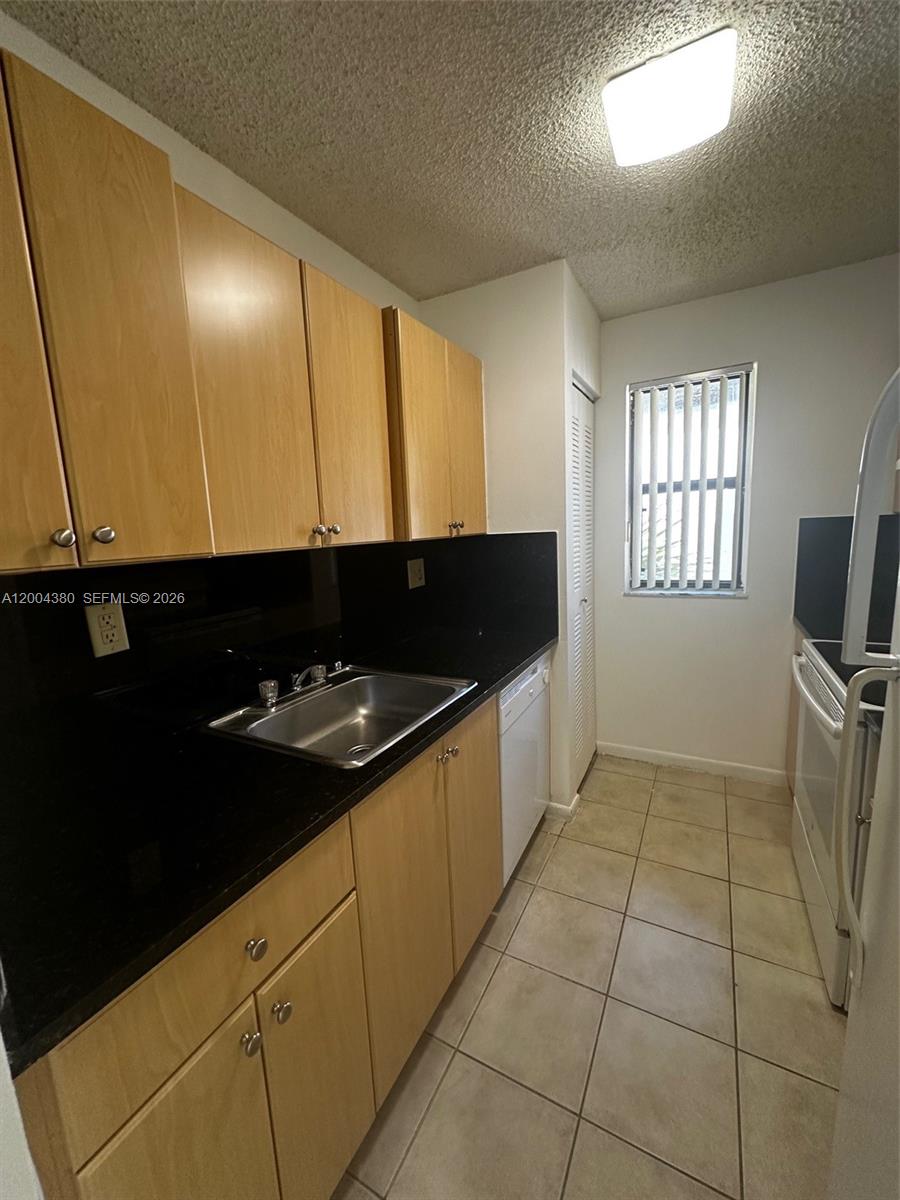 A galley kitchen features light wood cabinets, black countertops, a white dishwasher, and tiled floors — 7220 NW 179th St # 201, Hialeah FL 33015, HIALEAH, FL