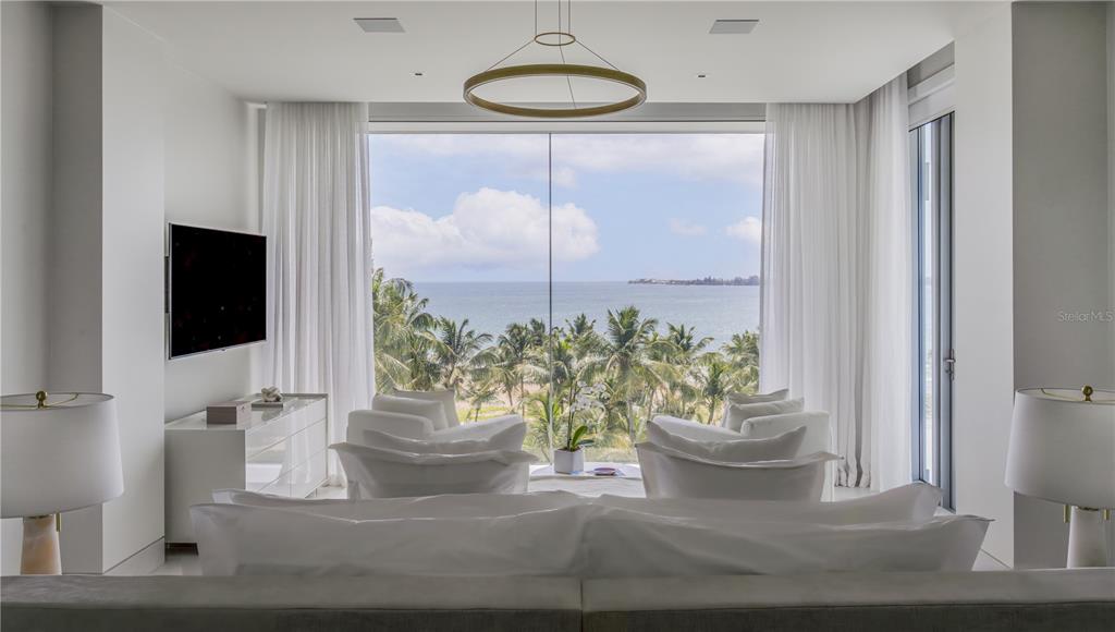 A modern bedroom features a large floor-to-ceiling window overlooking a beach with palm trees — Ocean Drive PH FOUR SEASONS PRIVATE RESIDENCES UNIT 6502, RIO GRANDE, PR