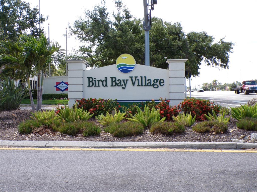 A sign for Bird Bay Village stands in a landscaped garden beside a road — 622 BIRD BAY DRIVE S UNIT 103, VENICE, FL