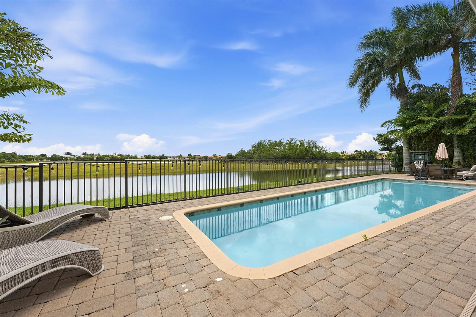 A rectangular swimming pool sits on a paved patio overlooking a lake and grassy field — 851 Edgebrook Lane, West Palm Beach FL 33411, WEST PALM BEACH, FL