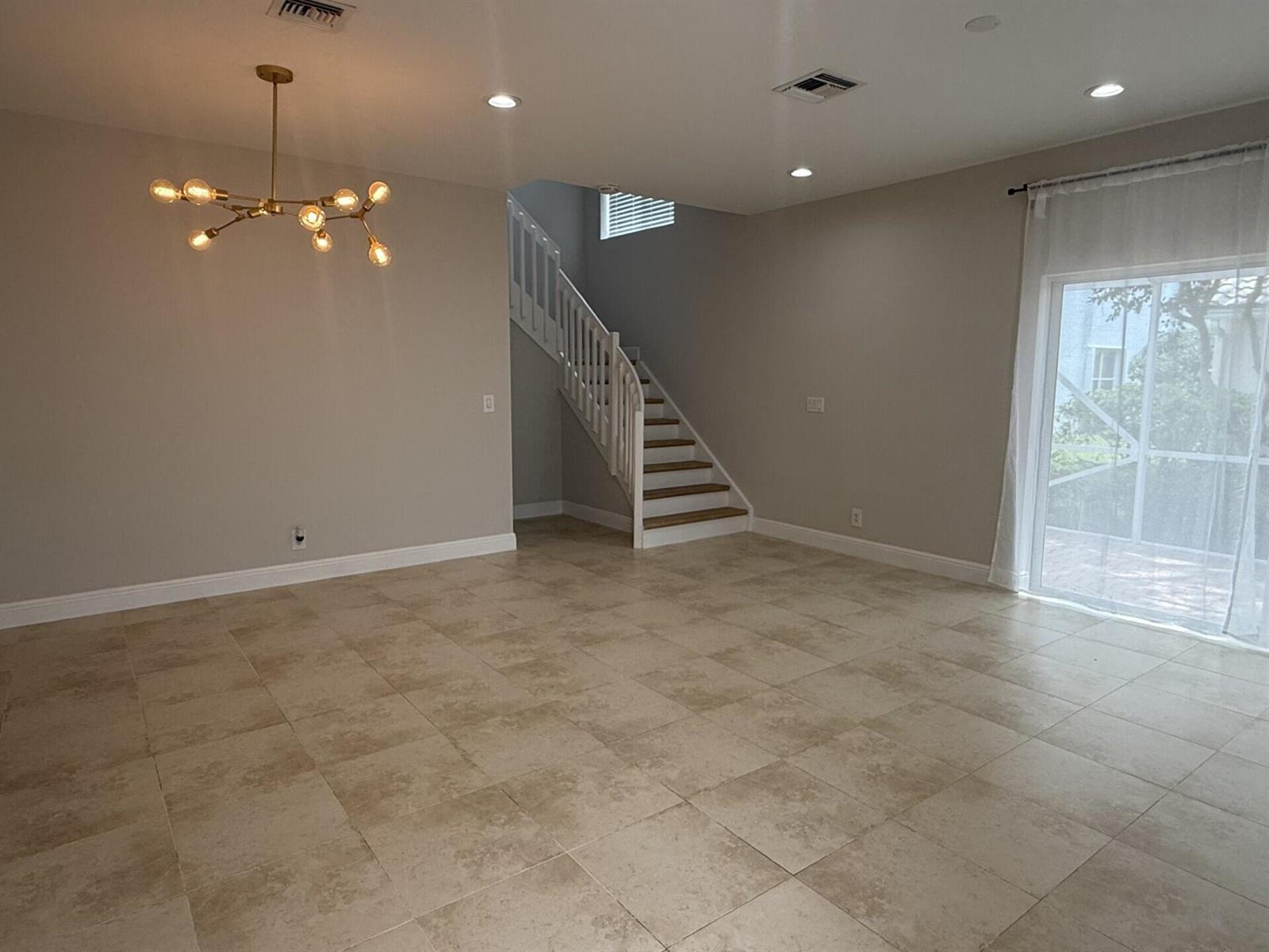 An open living area features beige tile flooring, a modern gold chandelier, and a staircase — 7218 Panache Way, Boca Raton FL 33433, BOCA RATON, FL
