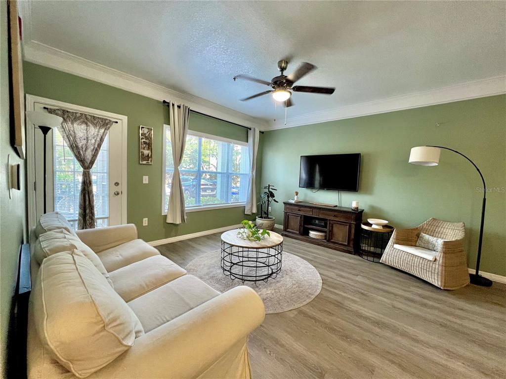 A living room features sage green walls, light wood flooring, a sofa, and a television — 4802 51ST STREET W UNIT 1315, BRADENTON, FL