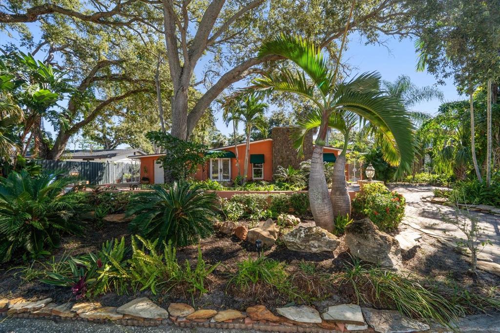 A single-story orange house with a stone chimney is surrounded by lush tropical landscaping — 3033 82ND WAY N, ST PETERSBURG, FL