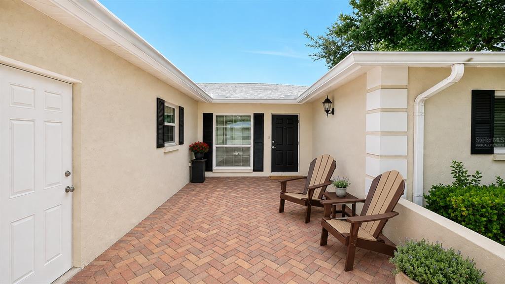 A paved courtyard features two wooden chairs, a black front door, and beige stucco walls — 5621 PALM AIRE DRIVE, SARASOTA, FL