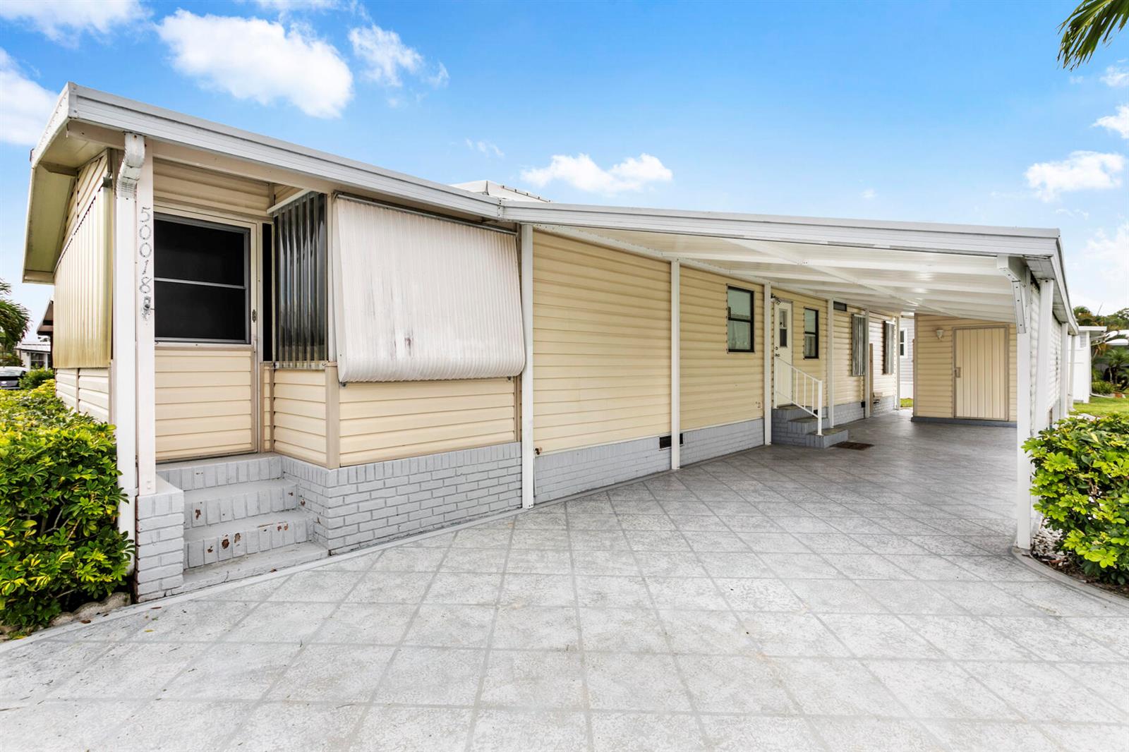 A light yellow mobile home features a covered carport, brick skirting, and a concrete driveway — 50018 Hacha Bay, Boynton Beach FL 33436, BOYNTON BEACH, FL