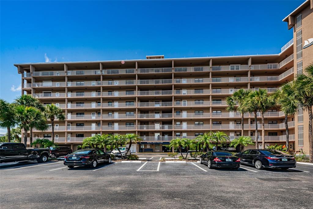 A multi-story apartment building with balconies stands behind a paved parking lot with cars — 19451 GULF BOULEVARD UNIT 301, INDIAN SHORES, FL