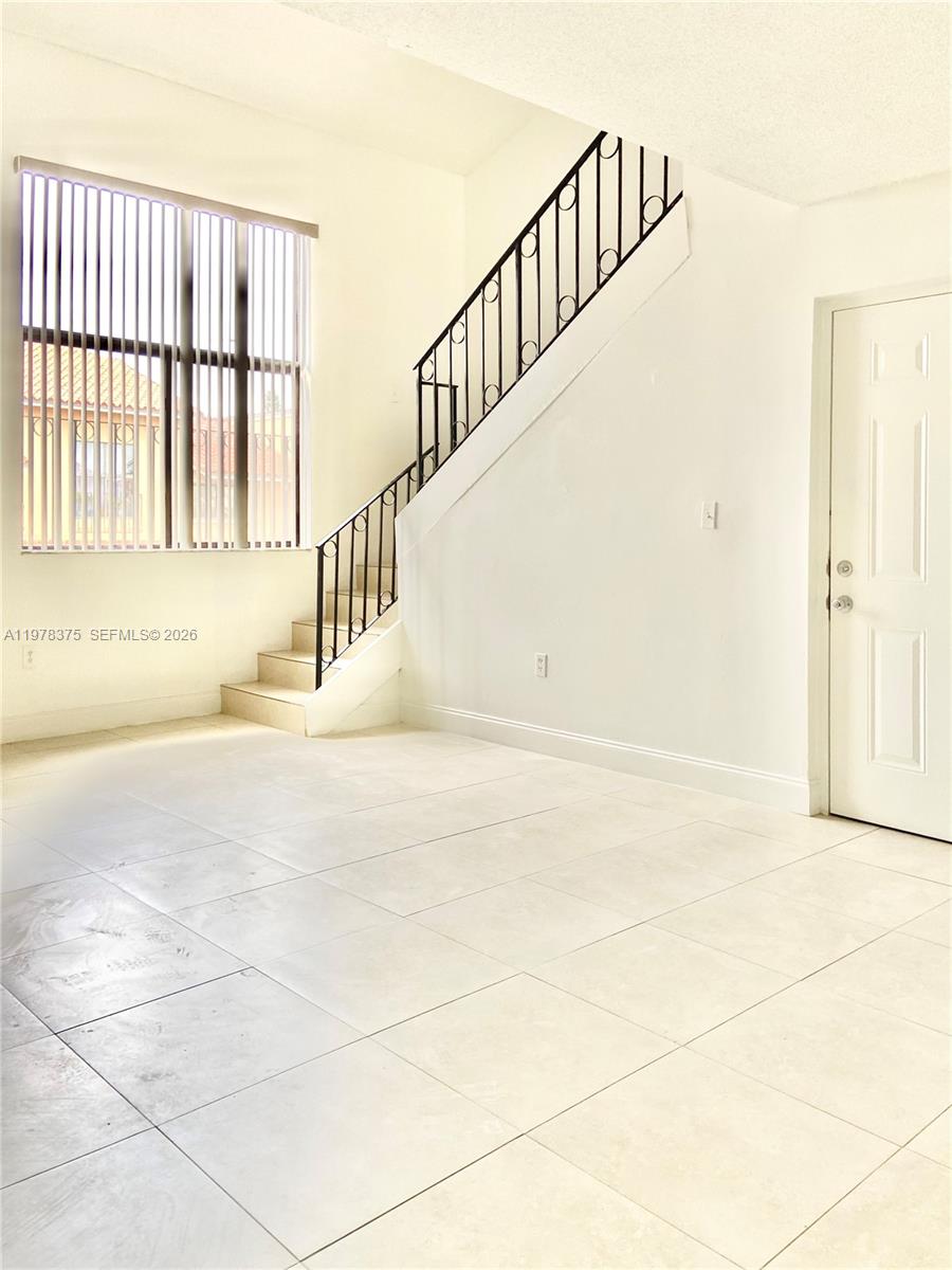 A bright living room features white tile floors, a staircase with black railings, and blinds — 2740 W 62nd St # 203, Hialeah FL 33016, HIALEAH, FL