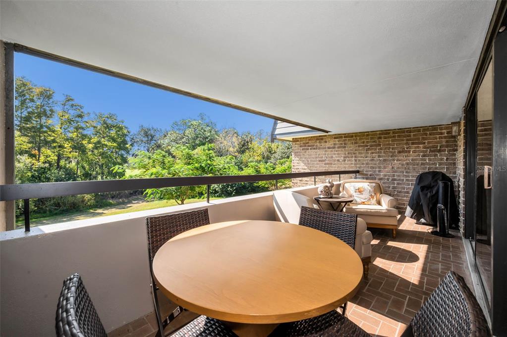 A covered balcony features a round wooden table, wicker chairs, and a view of trees — 36750 US HIGHWAY 19 N UNIT 28-2017, PALM HARBOR, FL