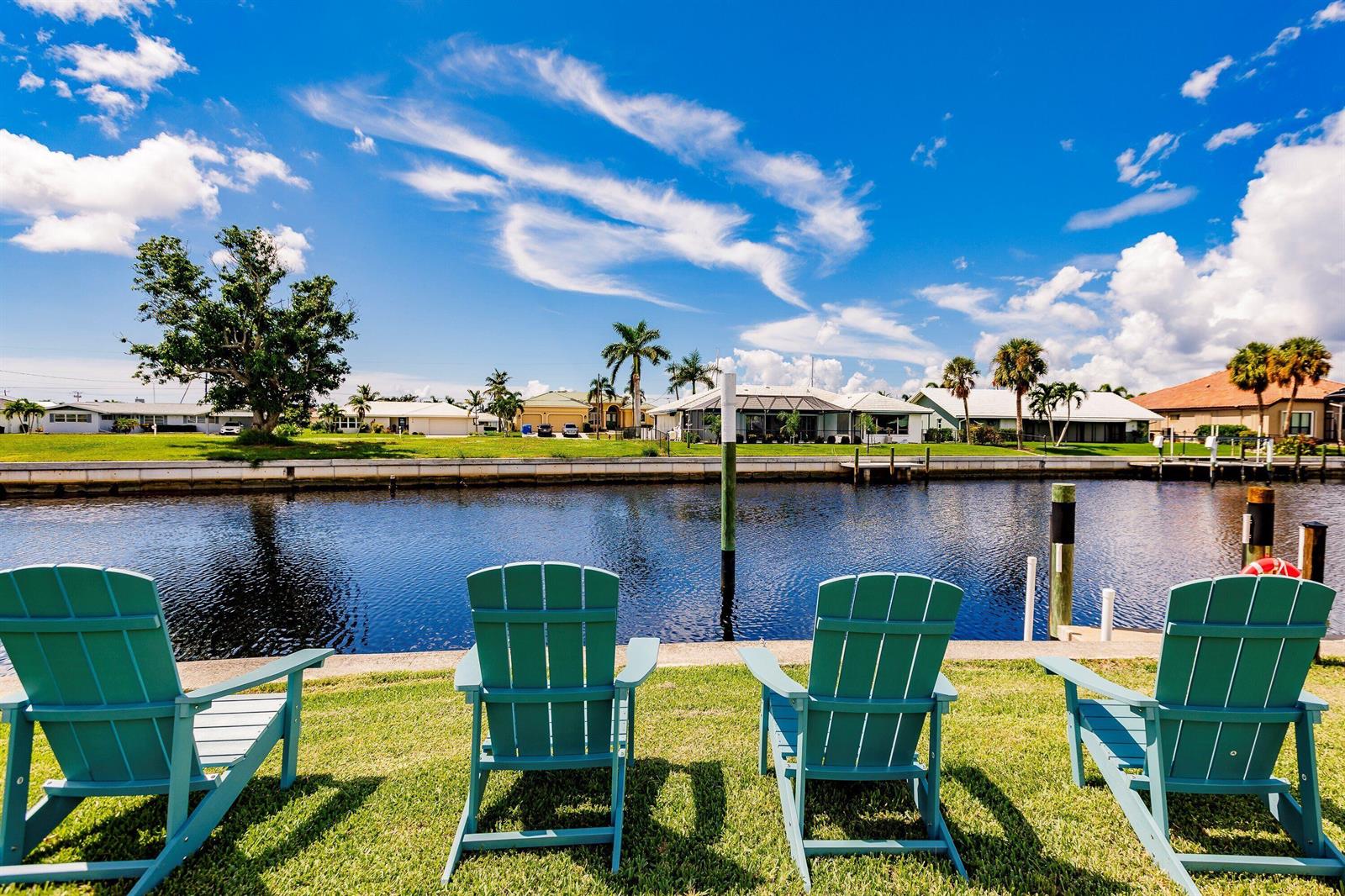 Four teal Adirondack chairs sit on a grassy lawn overlooking a canal and residential homes — 131 Casa Lane, Punta Gorda FL 33950, PUNTA GORDA, FL