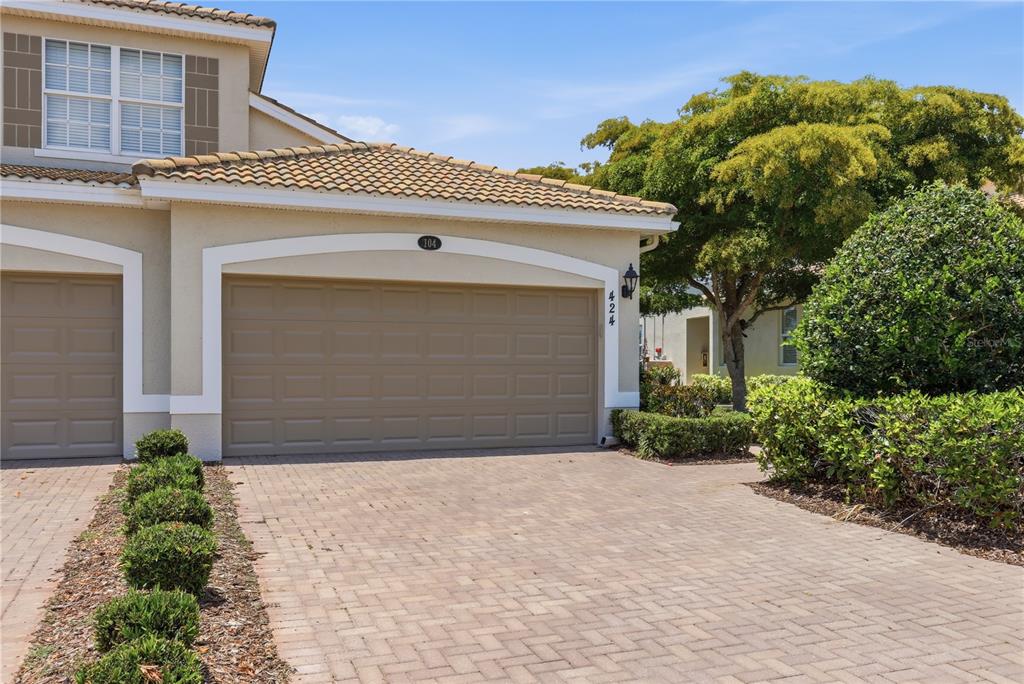 A beige house exterior features a two-car garage, a tiled roof, and a paved driveway — 424 WINDING BROOK LANE UNIT 104, BRADENTON, FL